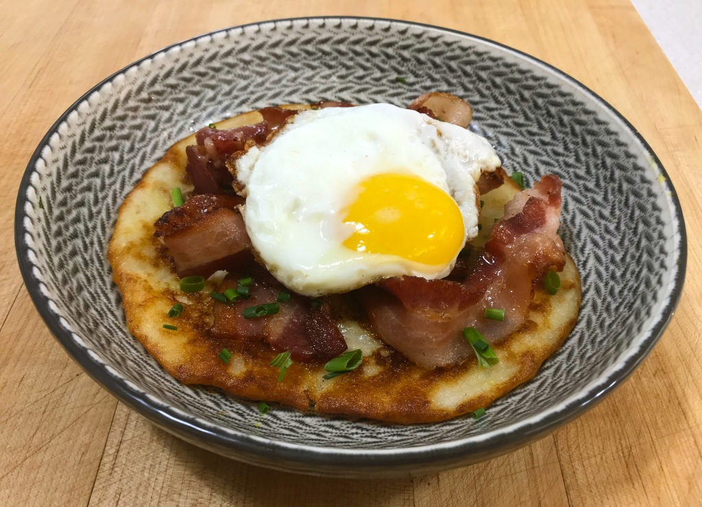 A fried egg, sitting on top of bacon, on top of a finished boxty pancake. Chopped chives are scattered around.