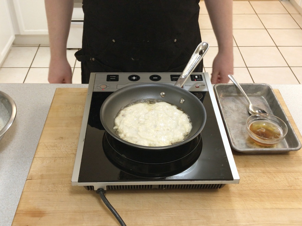 The boxty batter frying in bacon fat in a small non-stick frying pan. The bacon fat is bubbling around the sides.