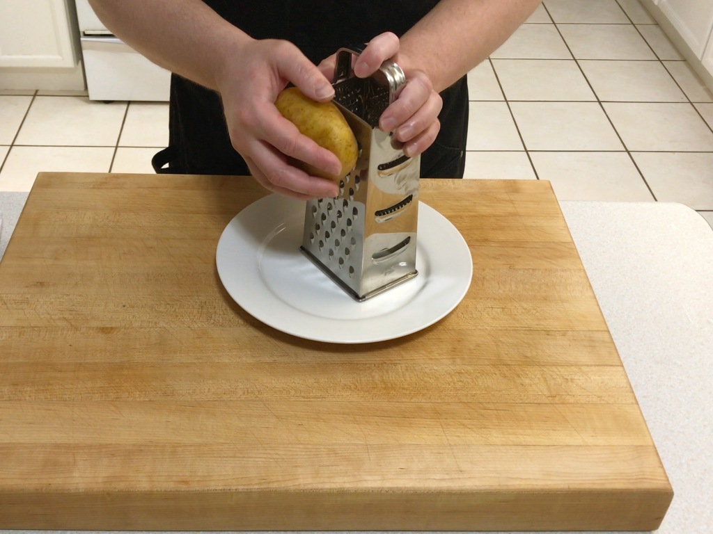 Grating a skin-on potato through the coarse holes on a box grater, onto a plate.