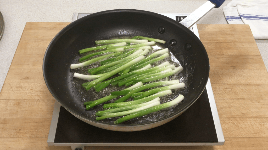 The green onions are browning in butter in a large non-stick pan. The tail end of the green onions has been chopped off, for use in other recipes, leaving the white and the first part of the green.