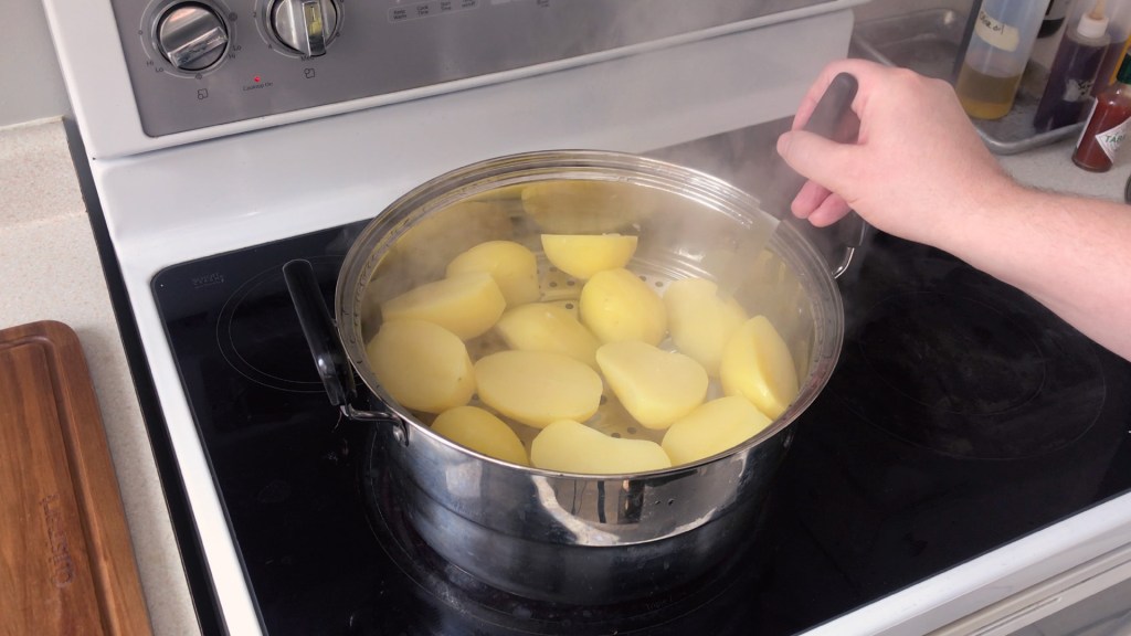 Peeled potatoes cut in half lengthways, in a steamer on the stovetop. A paring knife is being inserted into the potatoes to test if they are cooked.