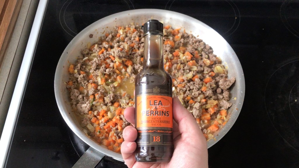 A bottle of Lea & Perrins Worcestershire sauce held above the pan containing the vegetables, meat and stock. The orange label is a common sight in the British pantry.